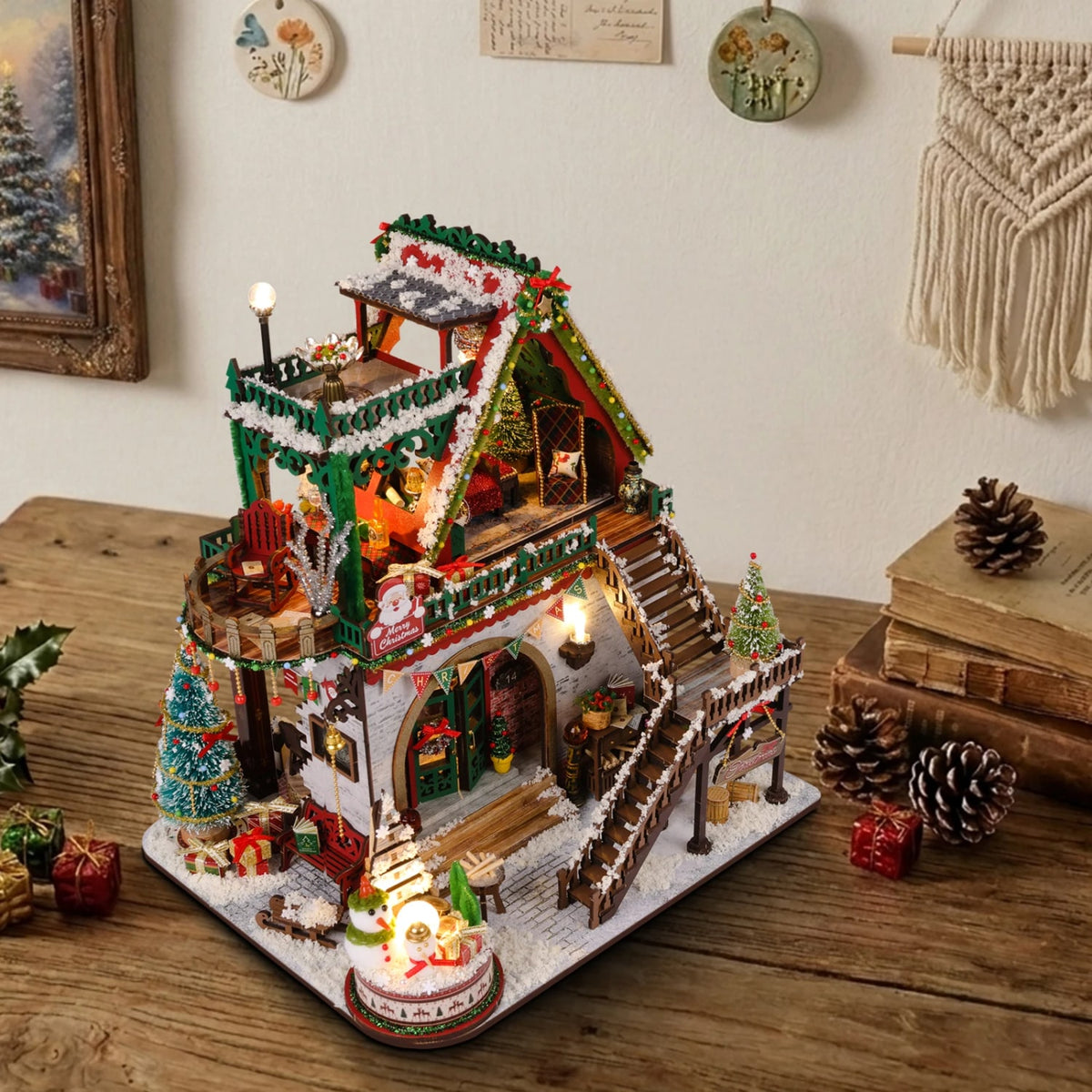 Miniature Christmas lodge kit glowing on a wooden table next to pine cones and old books