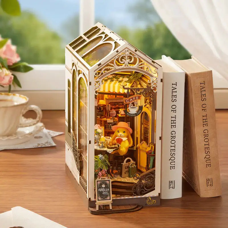 Holiday Cafe DIY book nook kit. Miniature wooden cafe diorama with warm interior, snowy window display, holiday lights, and steaming drinks. Designed to slide between books on a bookshelf.